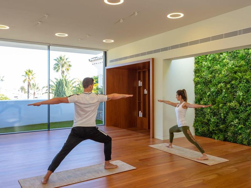 Twee personen doen yoga op matten in een zonovergoten ruimte met uitzicht op palmbomen en een groene muur.