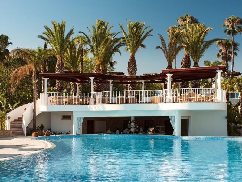 Moderne witte zwembadruimte met bar en palmbomen onder blauwe lucht.