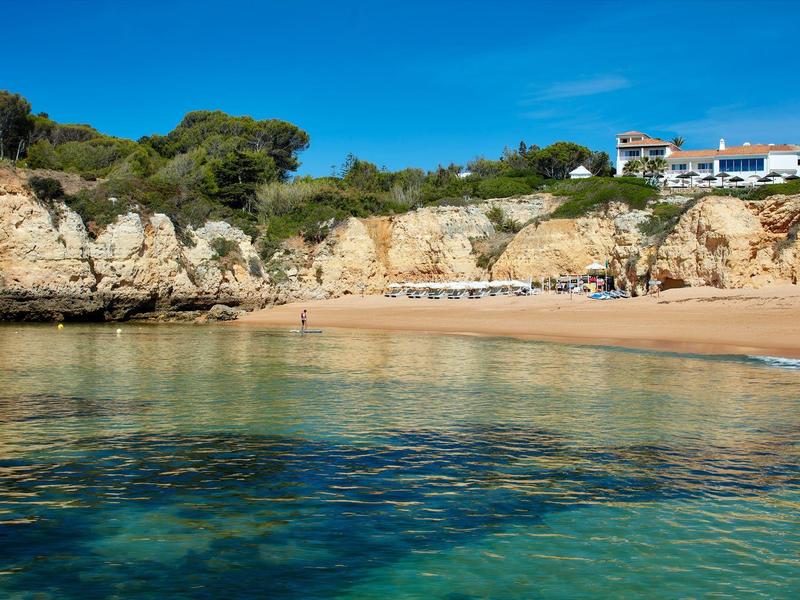 Helder zeewater bij een rustig zandstrand met rotsen en huizen op de achtergrond.