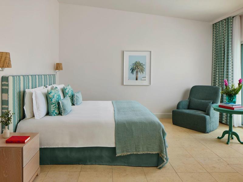 Lichte hotelkamer met groot bed, groene fauteuil, nachtkastje en minimale decoratie.