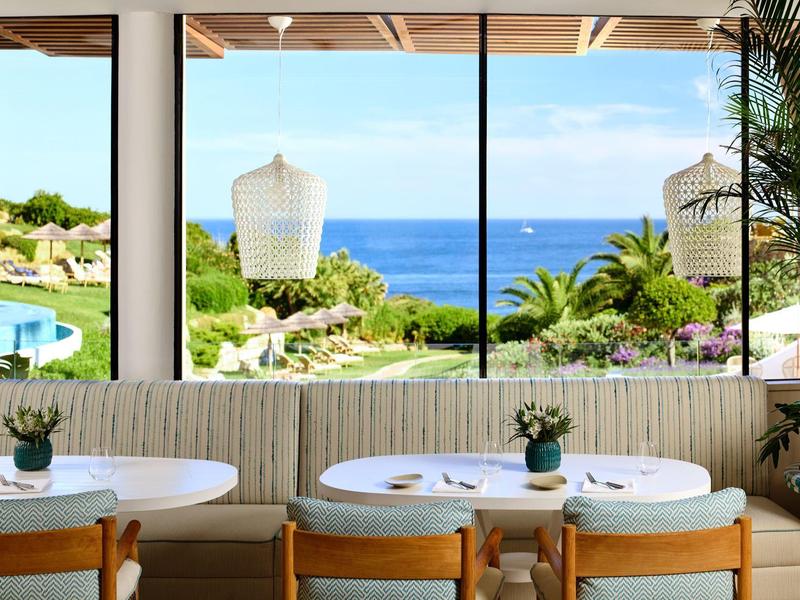 Eetgedeelte met houten stoelen, witte tafels en uitzicht op zee en tropische planten.