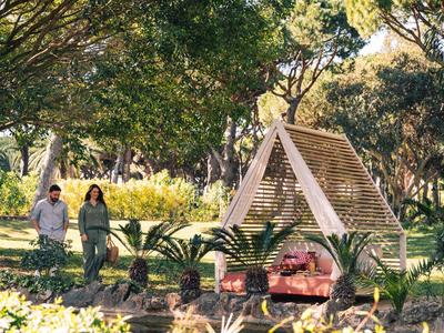 Paar spaziert neben einer hölzernen Gartenbank mit Dach in einem sonnenbeschienenen Park.