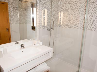 Moderne weiße Badezimmereinheit mit Waschbecken, Spiegel, Duschkabine und Musterfliesen an der Wand.