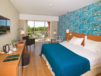 Helles Hotelzimmer mit großem Bett, blauem Bettüberwurf, Schreibtisch, Fernseher und Balkon mit Gartenblick.