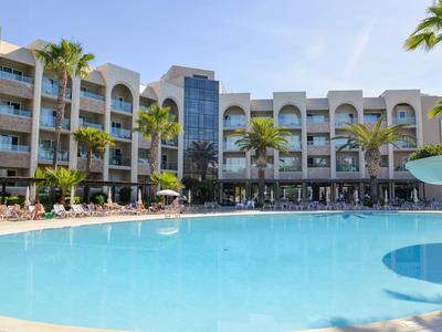 Großer blauer Pool vor hellem Hotelgebäude mit mehreren Stockwerken und Palmen.