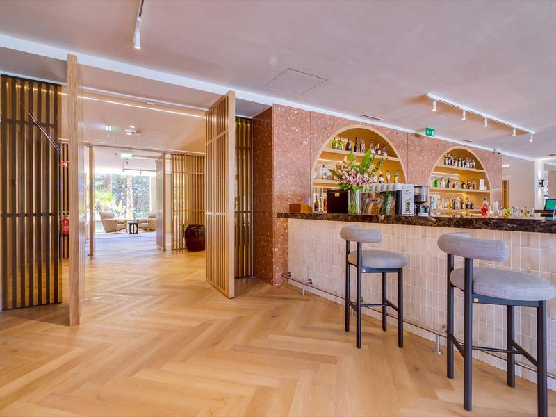 Moderní hotelový bar s dřevěnou podlahou, stoličkami a dekorativními nástěnnými policemi s rostlinami.