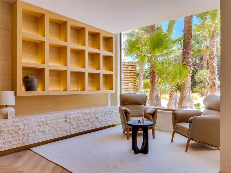 Jasná čekací zóna s moderními křesly, stolem a výhledem na palmy přes velká okna.