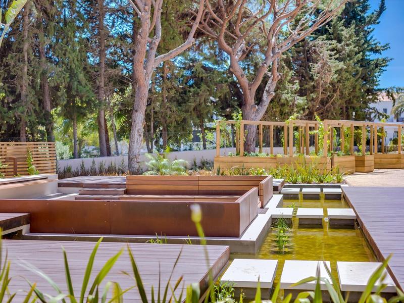 Moderní venkovní prostor s dřevěnou terasou, vodními plochami a zelení pod modrou oblohou