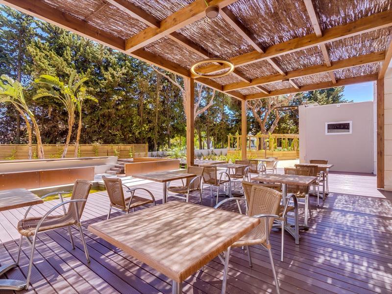 Zastřešená terasa s několika stoly a židlemi v zahradní oblasti.