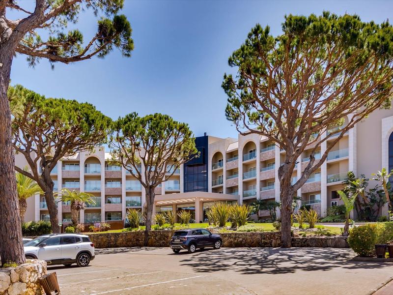 Moderní vícepatrová budova hotelu obklopená velkými upravenými stromy a zaparkovanými auty.