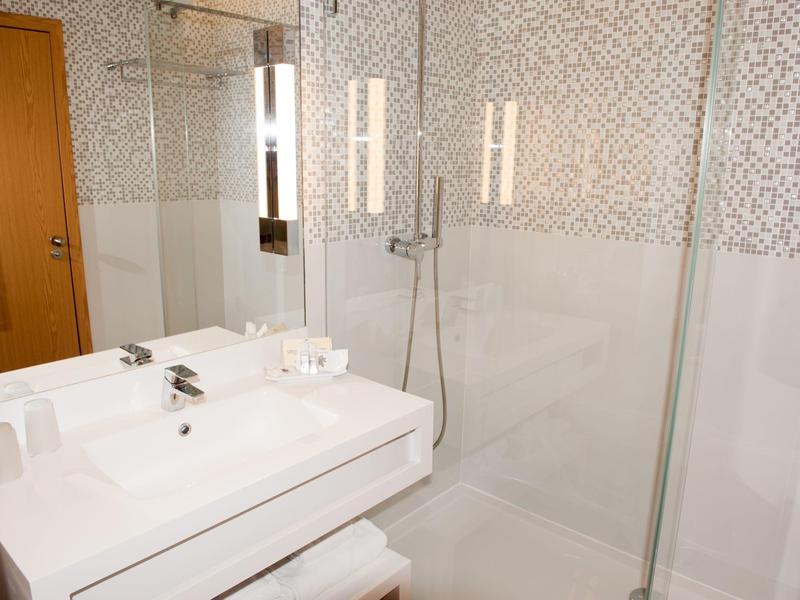 Moderní koupelna s bílým umyvadlem, skleněným sprchovým koutem a velkým zrcadlem.