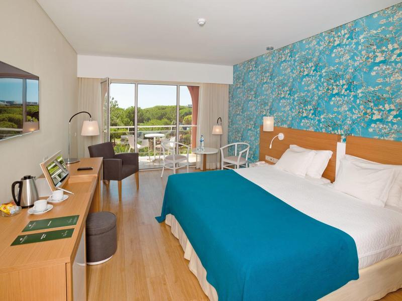 Moderní hotelový pokoj s velkou postelí, psacím stolem, modrým akcentem a balkonem s výhledem do zeleně.