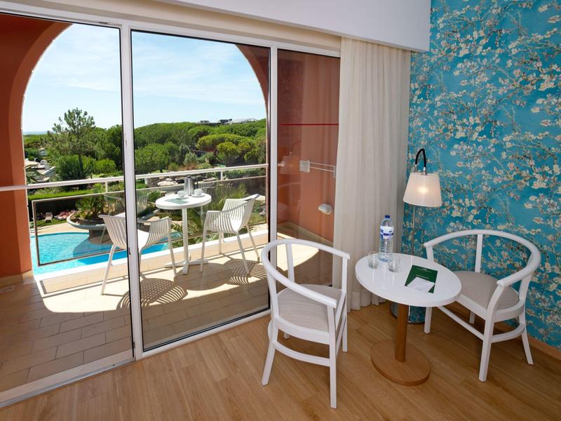 Světý hotelový pokoj s terasou, výhledem na bazén a zelenou krajinu.