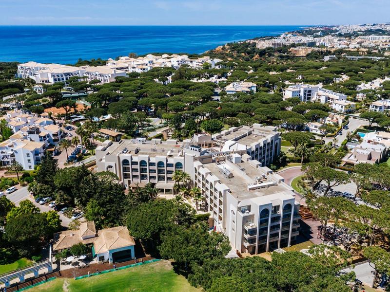 Letecký pohled na pobřežní hotel obklopený zelení s modrým mořem v pozadí.