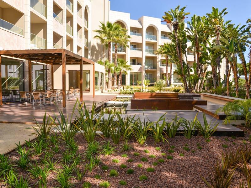 Hotel s venkovní částí, bazénem, palmami a sluneční terasou za jasné oblohy.