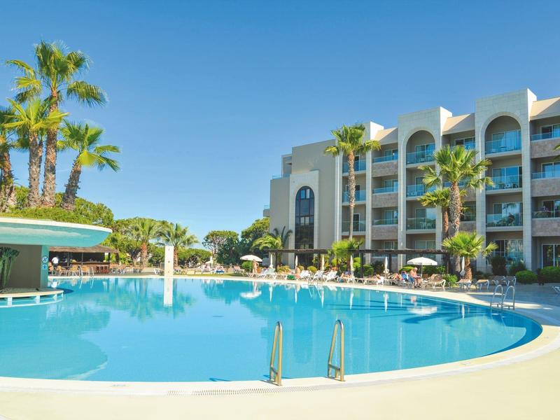 Moderní hotelový bazén s palmami a lehátky pod jasně modrou oblohou.