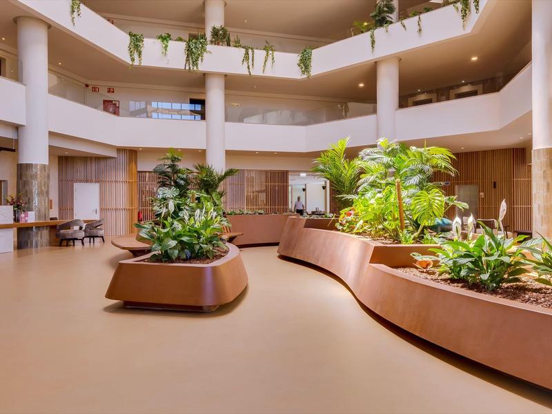 Světlá moderní hotelová hala s velkými rostlinami a zakřivenými lavicemi pod otevřeným atriem.