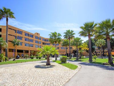Großes, gelbes Hotel mit Palmen und gepflastertem Weg unter blauem Himmel.