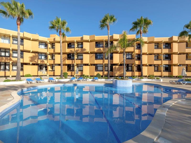 Ein großer Außenpool umgeben von drei- bis vierstöckigen gelben Wohngebäuden und Palmen.