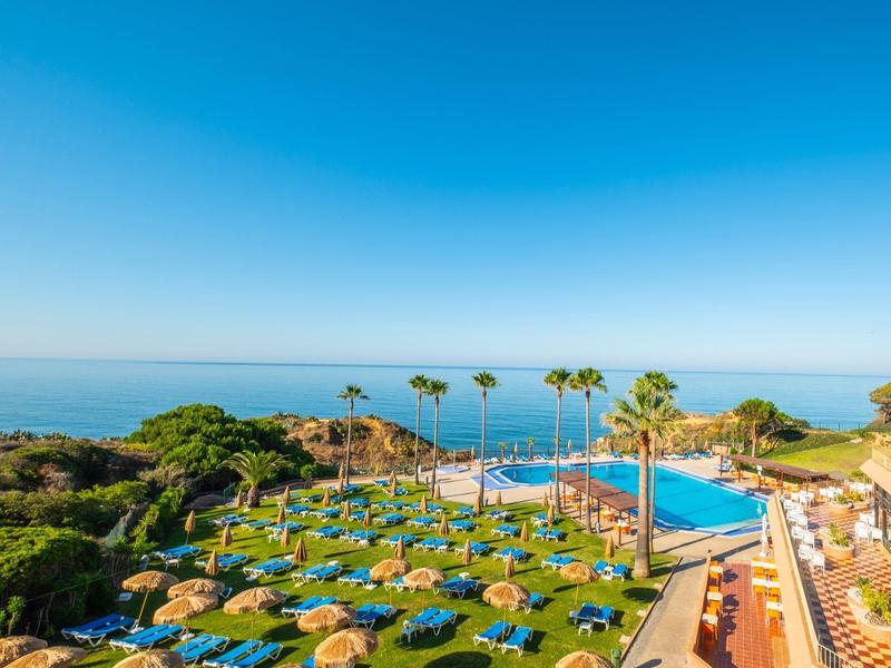 Strandhotel mit Pool, Palmen, Liegen und Blick auf ruhiges, blaues Meer bei klarem Himmel.