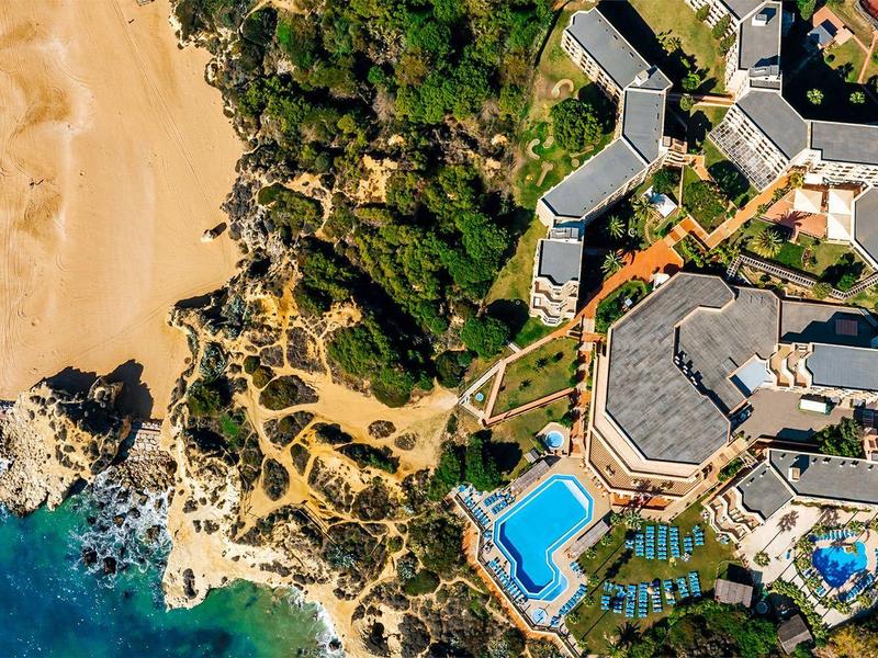 Luftaufnahme von Hotelanlage mit blauem Pool, grünen Bäumen und Sandstrand neben blauem Meer.