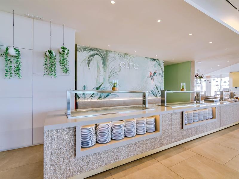 Moderne helle Cafeteria mit langer Theke, Tellerregal, Pflanzen und großem Wandbild.