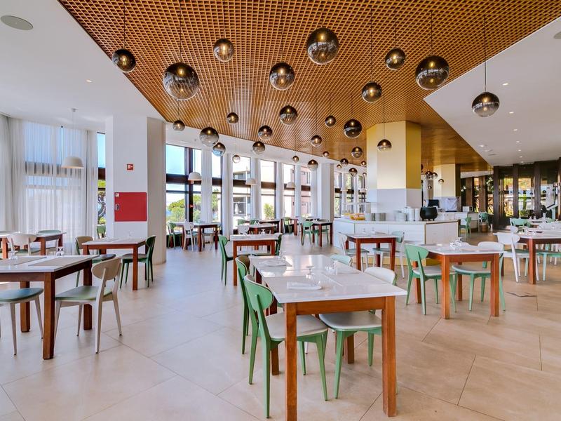 Moderner, heller Speisesaal mit Holztischen, grünen und braunen Stühlen, Kugelleuchten und großen Fenstern.