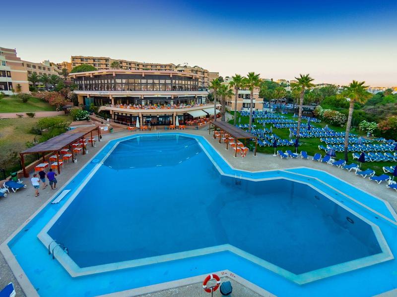 Großer blauer Pool mit Liegestühlen und mehrstöckigem Hotel im Hintergrund bei klarem Himmel.