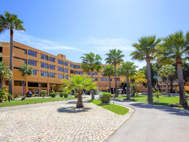 Großes, gelbes Hotel mit Palmen und gepflastertem Weg unter blauem Himmel.
