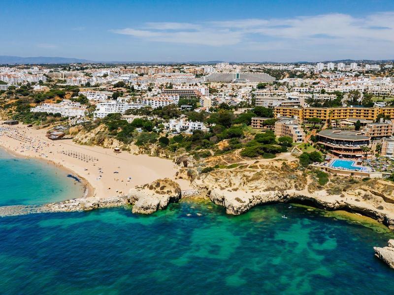 Strand mit türkisfarbenem Wasser, felsige Küste und Häuser im Hintergrund unter blauem Himmel.
