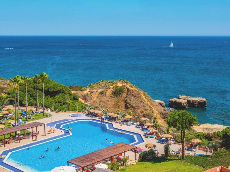Blick auf einen blauen Pool mit Sonnenschirmen, Palmen und Klippen am azurblauen Meer.