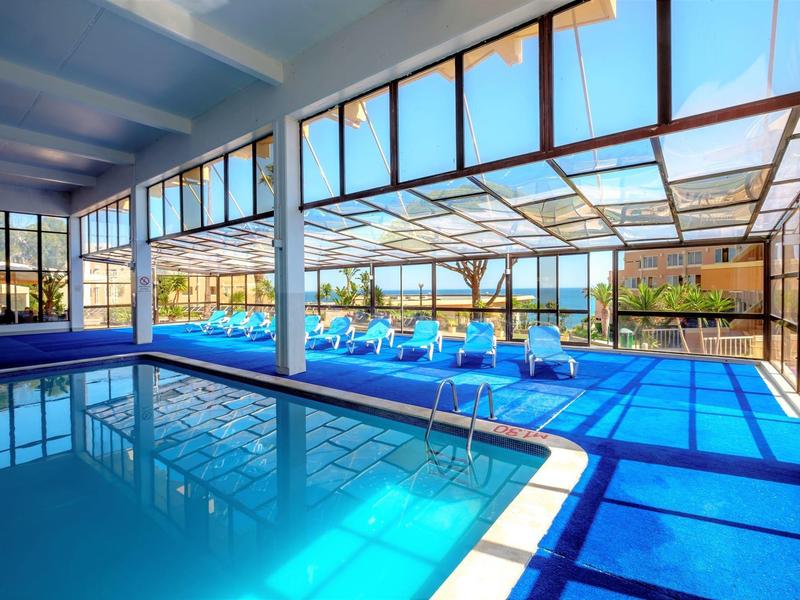 Innenraum mit großem rechteckigem Schwimmbecken, blauen Liegen und gläserner Dachverglasung.