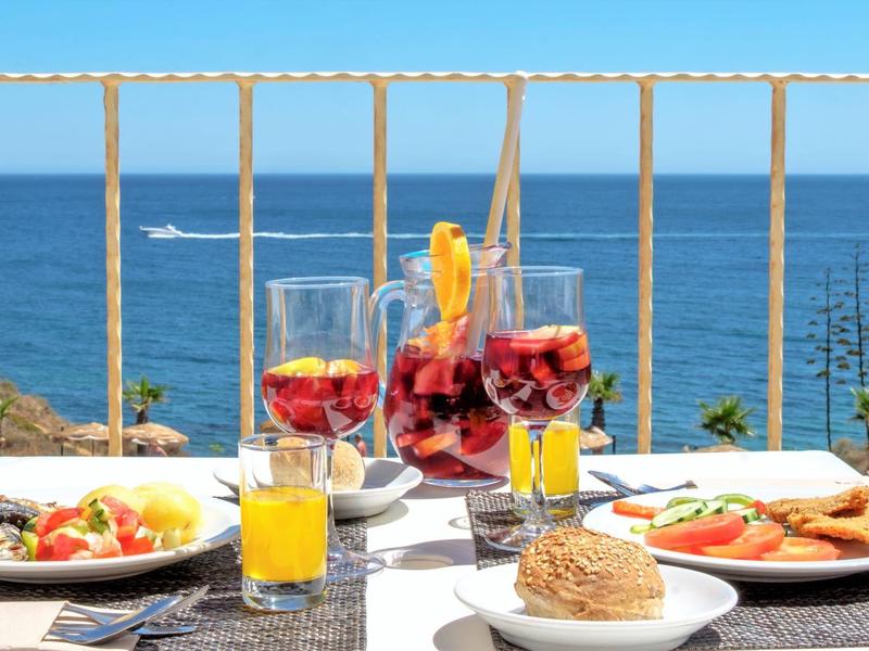 Tisch mit Getränken und Essen am Meer, im Hintergrund blaues Wasser und blauer Himmel.