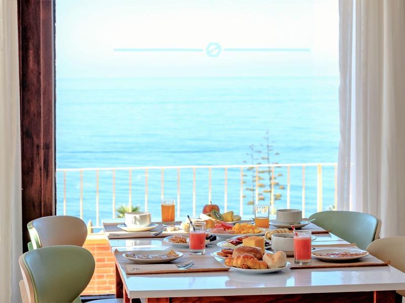 Esstisch mit Frühstück vor großem Fenster mit Blick auf das blaue Meer und hellen Himmel.