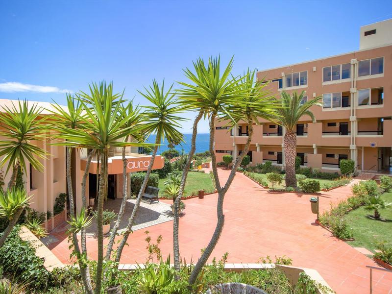 Innenhof mit Palmen, roten Gebäuden und Blick aufs blaue Meer unter klarem Himmel.