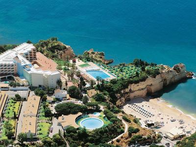 Luftaufnahme eines Küstenresorts mit mehreren Gebäuden, Pools, Gartenanlagen und Sandstrand am Meer.