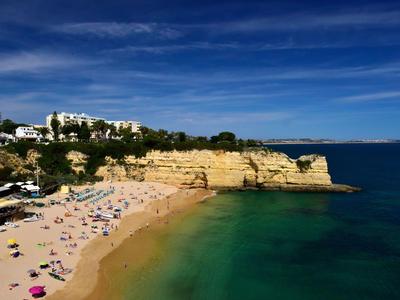 Spiaggia di sabbia con persone, scogliere rocciose e case bianche sotto un cielo azzurro