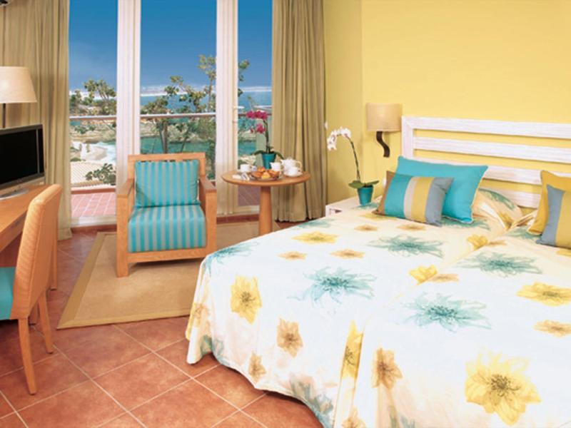 Camera d'albergo luminosa con letto matrimoniale, copriletto floreale, zona salotto e vista città.