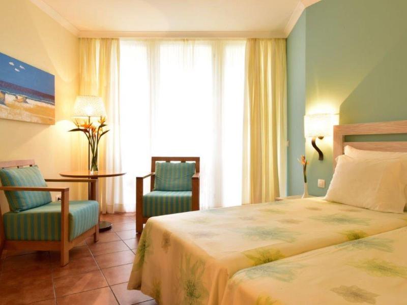 Camera d'hotel luminosa con grande finestra, letto matrimoniale e due poltrone imbottite accanto a un piccolo tavolo.