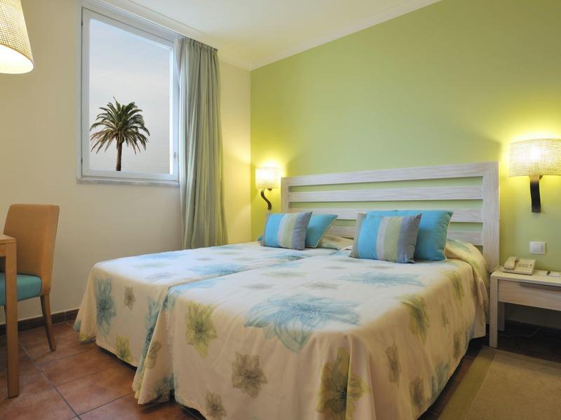 Camera d'hotel accogliente con letto matrimoniale, sedia, comodino e finestra con vista su palma.