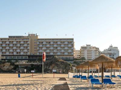 Plage avec parasols et transats devant un grand hôtel sous un ciel clair.