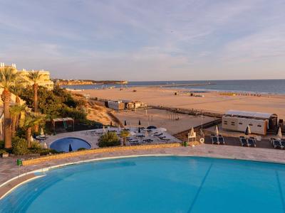 Vue sur piscine avec chaises longues et plage avec mer au coucher du soleil.