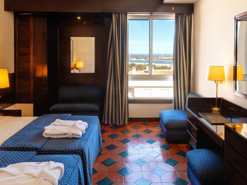 Chambre d'hôtel bien éclairée avec deux lits simples, couettes bleues et vue sur la ville depuis la fenêtre.