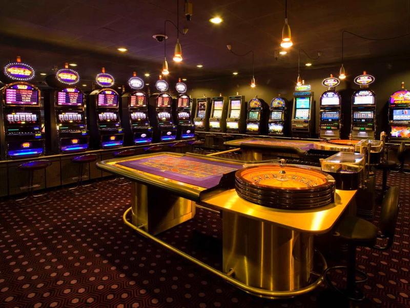 Vue d'une salle de jeux de casino avec machines à sous, table de roulette et éclairage tamisé.