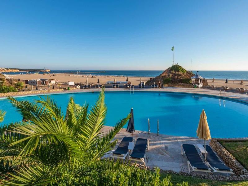 Piscine ronde avec eau bleue, entourée de chaises longues, vue sur la mer et ciel bleu clair.