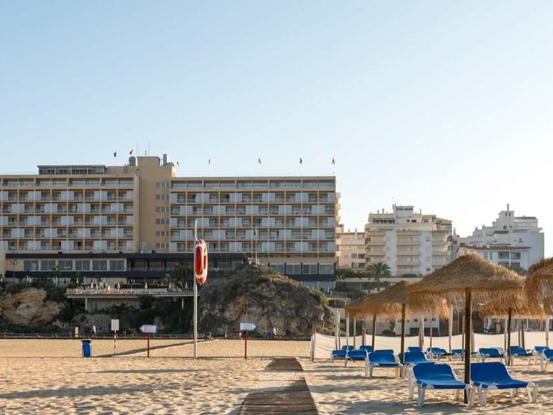 Plage avec parasols et transats devant un grand hôtel sous un ciel clair.