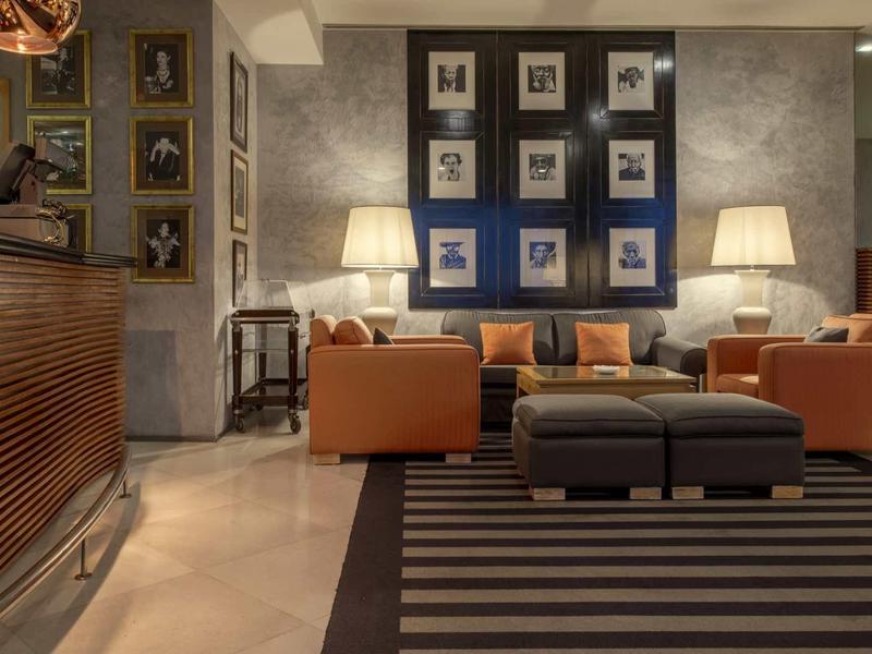 Un espace lounge d'hôtel élégamment aménagé avec des fauteuils, des canapés, des lampes et un mur décoré de tableaux.