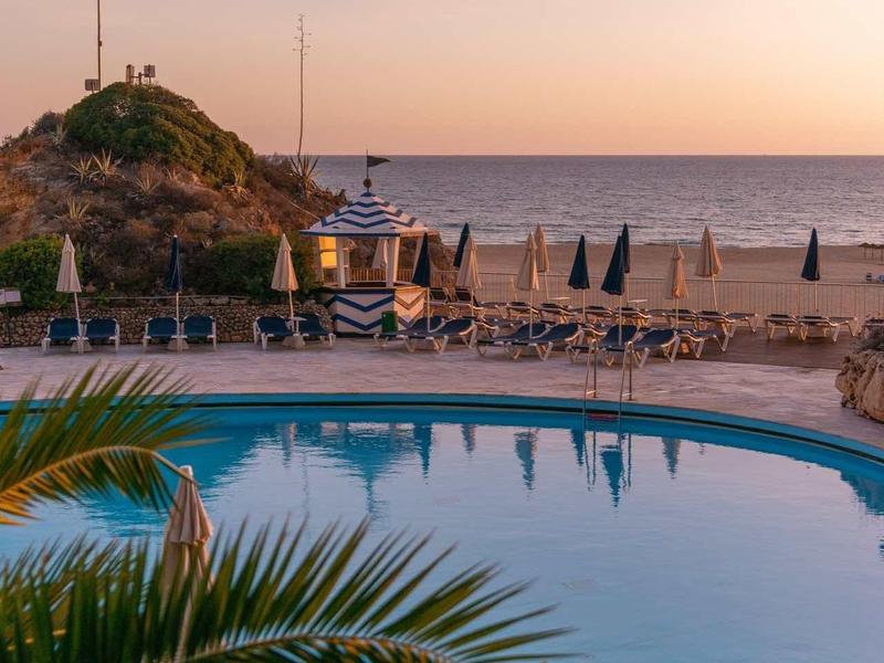 Piscine ronde près d'une plage avec espaces détente et vue sur la mer au coucher du soleil.