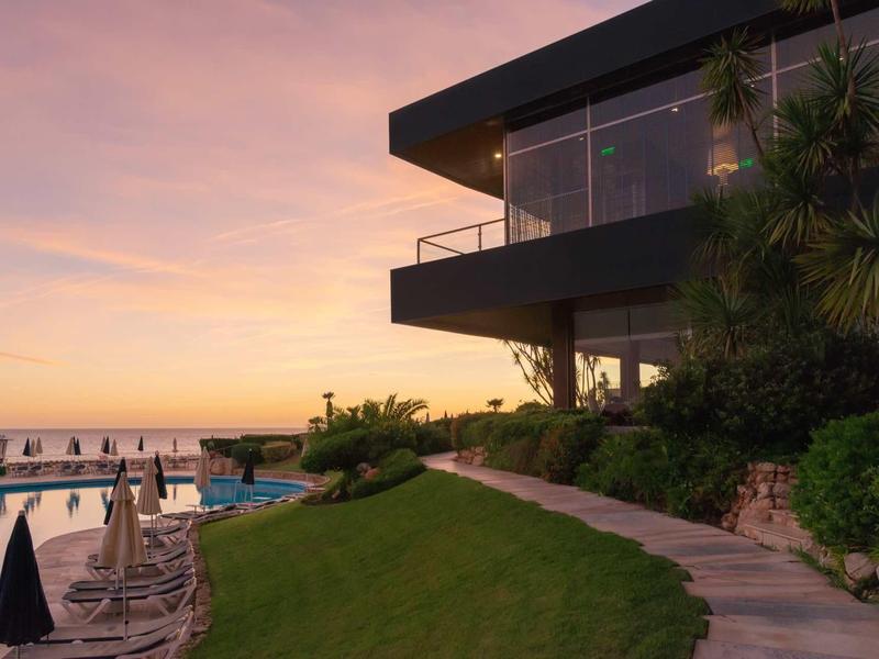 Hôtel moderne avec piscine et terrasse au coucher du soleil donnant sur la mer.