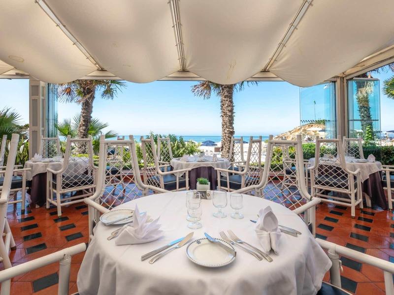 Terrasse de restaurant avec chaises et tables blanches, vue sur des palmiers et la mer.